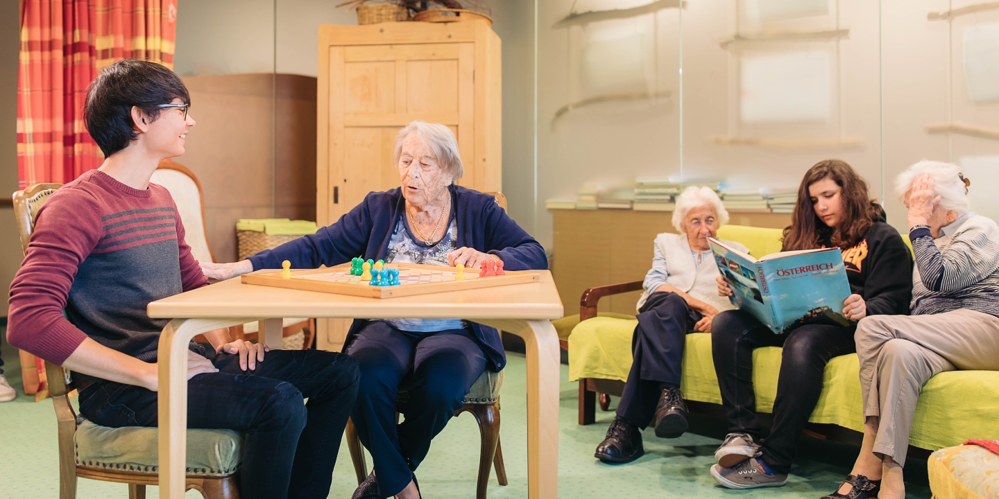 Jugendliche lesen Senior*innen ein Buch vor und spielen ein Brettspiel mit ihnen.