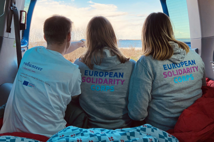 Drei junge Erwachsene sitzen hintenin einem Van und schauen zur Tür hinaus und haben Pullover auf auf denen steht Euorpean solidarity Corps