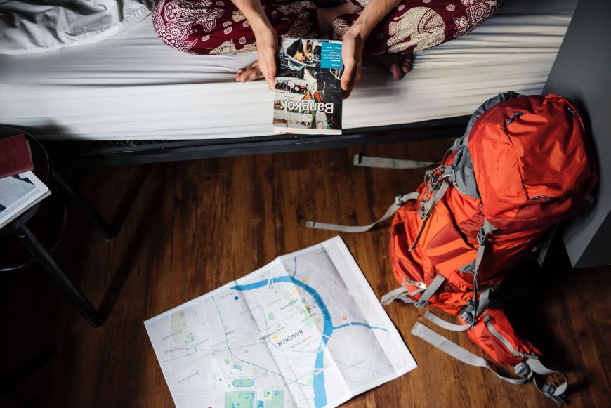 Eine Person sitzt mit einen Reiseführer über Bangkok auf dem Bett, am Boden steht ein roter Rucksack und liegt eine Landkarte.
