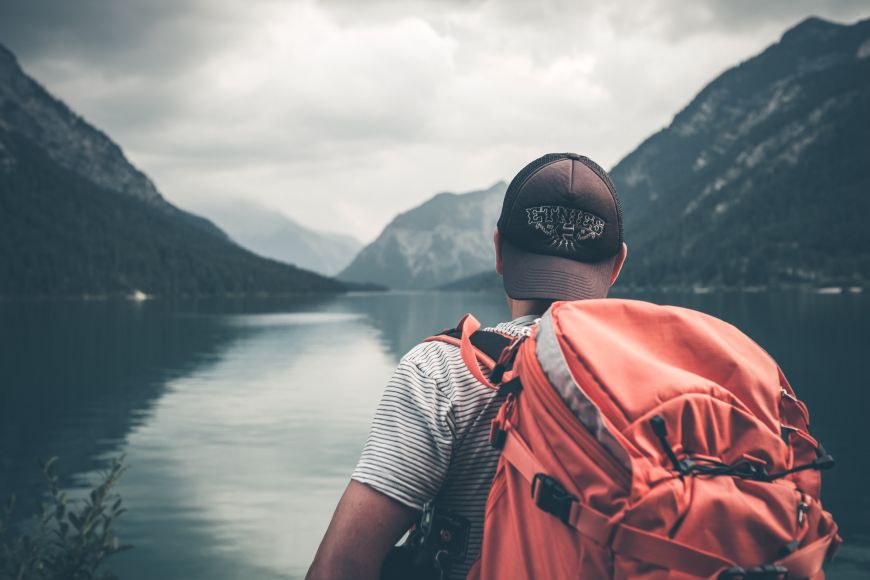 Junger man steht mit einem roten Rucksack bei einem Bergsee