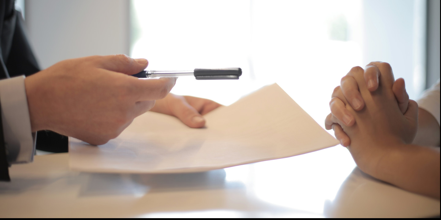 Eine Person reicht einer anderen Person einen Stift und Unterlagen zur Unterschrift.