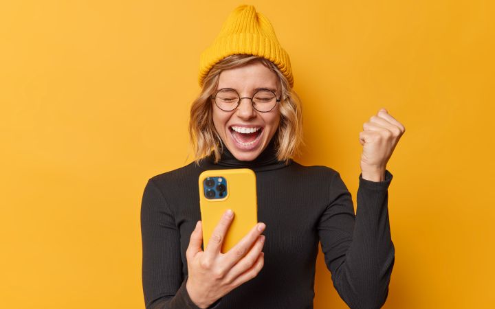 Eine junge frau mit gelber Kappe und Handy mit gleber Hülle ballt die Faust vor freude vor einer gelben Wand.